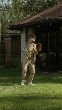 Full vertical slowmo of happy teenage African American boy playing baseball outdoors with friend or family on summer day, on green lawn, throwing ball and catching it using leather glove