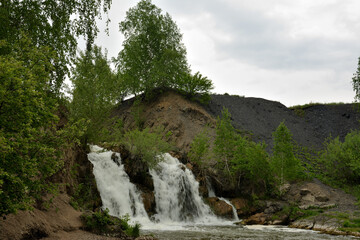 Obraz premium The forked river bed flows like a stormy waterfall from a high rocky cliff on a cloudy summer day, surrounded by tall birches.