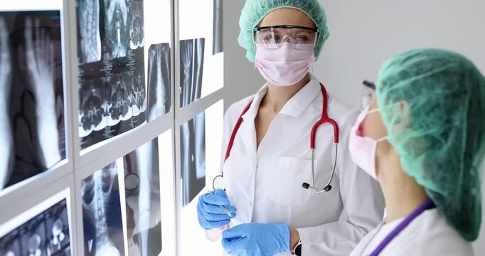 Woman doctor in mask attaches X-ray image on light display with assistant in hospital. Medical specialists check patient radiograms in clinic