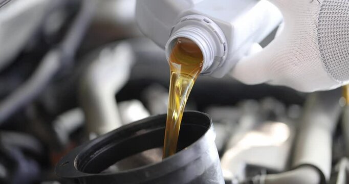 Pouring golden-brown motor oil from plastic bottle into engine of vehicle through black funnel. Process of adding oil to car engine for maintenance