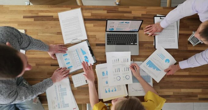 Colleagues discuss business data studying charts at desk in office upper view. Coworkers brainstorm ideas for marketing strategy in boardroom