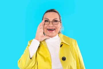 Woman in yellow coat smiles and gestures enthusiastically against a bright blue background during a cheerful moment