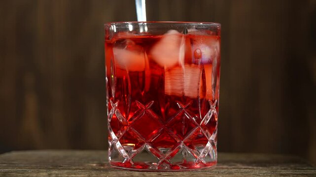 Cocktail spoon stirring a red drink in a glass. Red cocktail in crystal glass on wooden table, close up. Summer ice drink - tea or juice with ice cubes