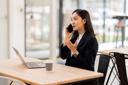 Happy young business woman manager talking consulting client on cell phone working on laptop in office. Busy woman professional business making call on cell using computer sitting at desk
