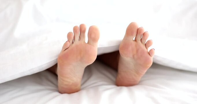 Pair of person bare feet visible under white blanket and toe scratches sole. Scene suggests relaxation in bed with snow-white bedding in early morning