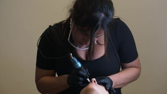 With deliberate control, esthetician leans over client, guiding the microblading tool with both hands. Her posture is firm, eyes fixed on the brow line, capturing the intensity of real-time precision.