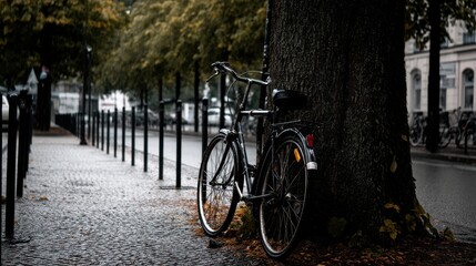 Fototapeta premium Black bicycle leaning against a tree on a rainy urban street with cobblestone pavement, surrounded by trees and bike stands under gray clouds