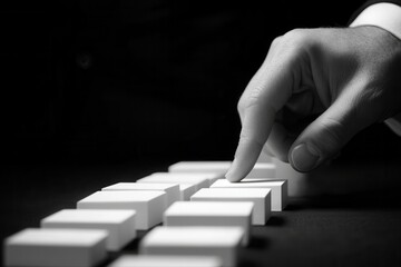 Hand poised to topple a line of white dominoes in a dramatic black and white setting