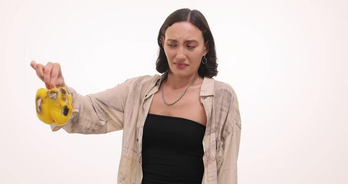 Woman holds rotten bell pepper covered with green mold against white background. Female looks at spoiled vegetable with disgusted expression on face