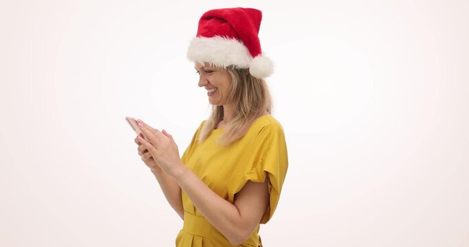 Woman smiles warmly as types holiday wishes on phone against white background. Female in red Christmas hat writes heartfelt message to best friend - Powered by Adobe