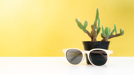 Stylish sunglasses rests on a white table, with a cactus plant in the background, showcasing a modern and minimalist aesthetic. isolated yellow background and copy space