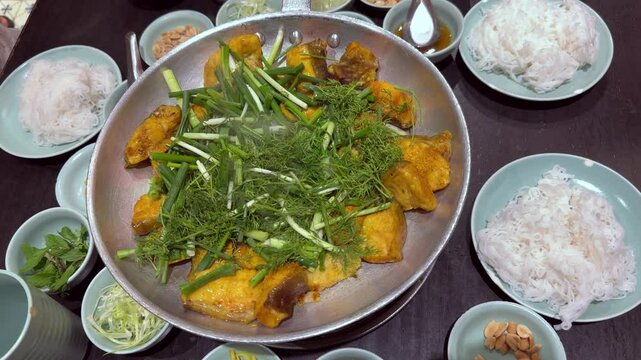 A dish of boneless grilled hemibagrus fish, a type of river catfish fried called Cha Ca La Vong Hanoi, table of diners with fresh dill and spring onion, eating with cilantro
