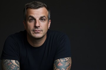 Fototapeta premium Tattooed man seated comfortably looks directly into the camera, showcasing intricate arm designs while creating a strong, confident presence in a dark setting