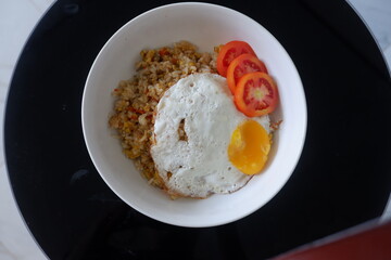 Delicious spicy village-style fried rice served in a white bowl, topped with a sunny side up egg and accompanied by sliced fresh tomatoes.