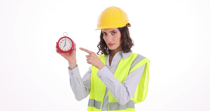 Forewoman in helmet shows alarm clock threatening by finger on white background. Female taskmaster reminds about deadline at construction site
