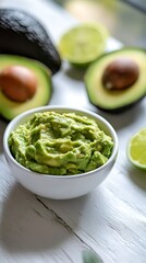 Obraz premium Creamy Green Guacamole Dip in Bowl with Fresh Avocados and Limes on White Surface