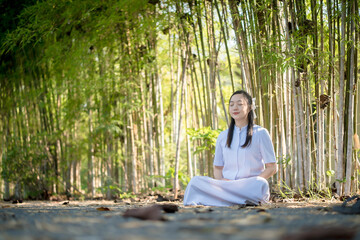 Young Asian woman peacefully meditating in a serene bamboo forest, eyes closed, embracing...