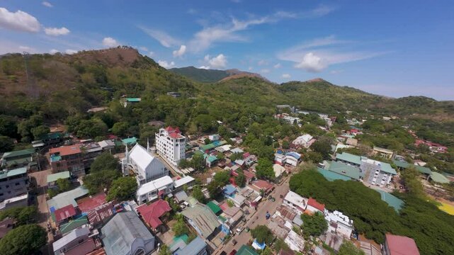 Quick daytime drone flyover of Coron town center in Palawan. The view captures rooftops, roads, green hills, cell towers, Mount Tapyas, and distant islands under a partly cloudy sky.
