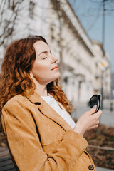 Young woman with auburn curly hair peacefully basking in the sun on a city sidewalk. City life...