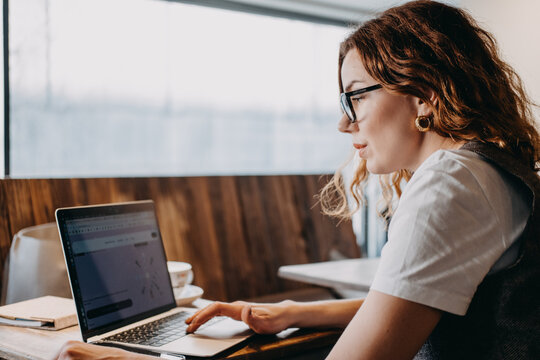A young woman with curly red hair concentrates on her laptop in a softly lit cafe. Focused productivity, deep work sessions, digital workflow, cognitive engagement...