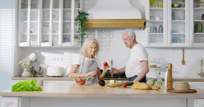 Positive old man and woman listening to music and dancing in kitchen, portrait. Good mood and emotions, have fun together, retirees feeling good and enjoying life, happy elderly spouses moving funny