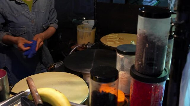 Night market vendor cleaning and preparing hot plate for making crepe, with various ingredients like bananas, chocolate chips, and colorful sprinkles readily available