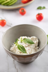 Gray bowl with cottage cheese, fresh basil and pepper. Light background with blurred cherry tomatoes and avocado. Concept of healthy and natural food