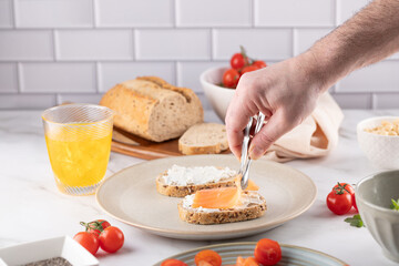 Male Hands Preparing Smoked Salmon Avocado Toast. Fresh Ingredients on Marble..Close-up of hands using tongs, kombucha, and cherry tomatoes. Authentic vibe for home chefs or healthy recipe blogs