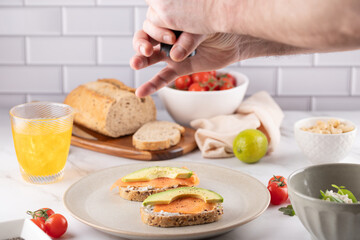 Healthy Living Concept: Adding Pepper to Salmon Avocado Toast. Marble table with kombucha and rustic bread. Great for meal kits or mindful eating campaigns