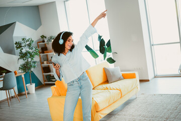 Young cheerful woman enjoying music and dancing in a colorful modern living room with headphones,...
