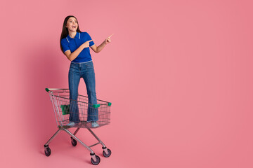 Young woman standing with gesture on shopping cart against pink background, promoting sale and...