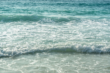 Close-up texture of ocean waves and sandy beach under sunlight with turquoise tones and soft foam