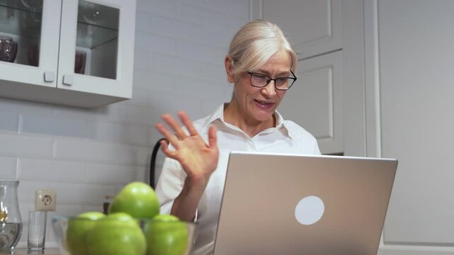 Elderly woman with glasses using a laptop in a modern kitchen with green apples, symbolizing remote work and healthy living.