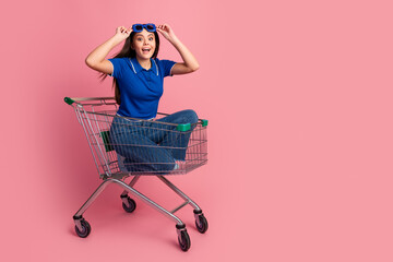 Joyful young woman posing playfully in a shopping cart against a vibrant pink background