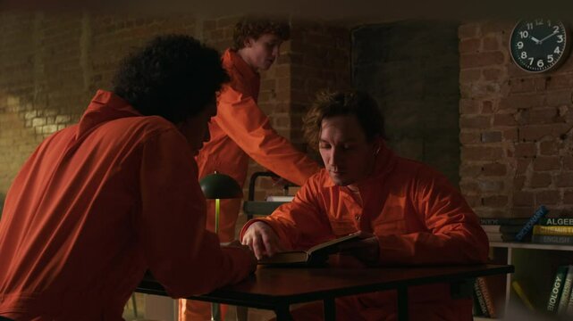 Two multiethnic male convicts in orange uniform discussing book at wooden table, young man with wheeled trolley giving out literature in prison library