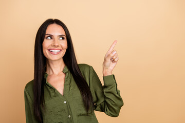 Smiling young woman in green blouse pointing to the side against beige background expressing enthusiasm and positivity