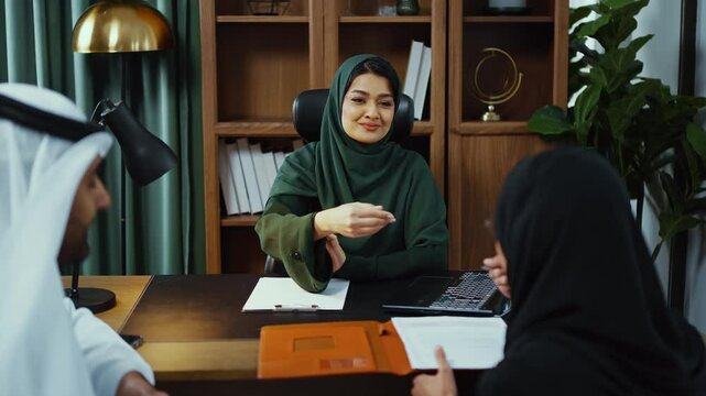 Emirati businesswoman in traditional attire leads a meeting with two female colleagues, gesturing and engaging in conversation across a desk in a modern office setting.