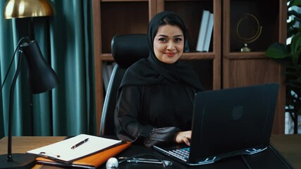 Young muslim doctor woman wearing traditional headscarf sitting at desk in office making video call with patient explaining diagnosis and writing notes on clipboard.