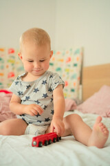 Naklejka premium Cheerful baby in star-patterned romper joyfully plays with a red toy train on a bed. The child exhibits excitement and curiosity, enhancing a playful and warm atmosphere.