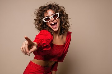 Trendy young woman with curly hair laughing joyfully, wearing bold red fashion outfit and white retro cat-eye sunglasses.
