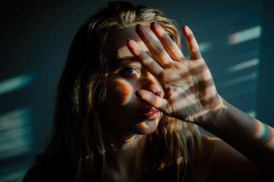 Introspective Young Woman Shielding Her Face from Light with Colorful Rainbow Reflections in Soft Blue Tones - Powered by Adobe