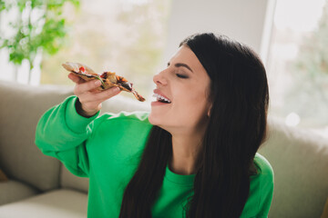 Young woman in a stylish green pullover enjoying a slice of pizza in a bright and cozy living room setting.