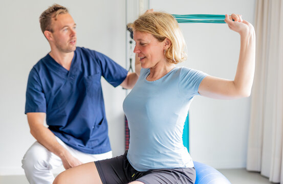 Physiotherapist assisting patient exercising with resistance band on fitness ball. Patient doing exercises with resistance band assisted by physiotherapist for rehabilitation