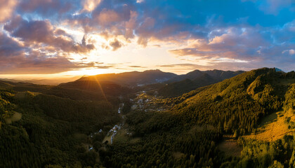breathtaking panoramic drone shot captures mountain range during sunset, with vibrant sunbeams breaking through clouds, illuminating lush green landscape below