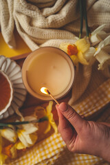 Close-up of a hand lighting a candle with a match, surrounded by daffodils, a knit blanket, and yellow checkered fabric in a warm and cozy springtime setting
