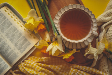 Flat lay with an open Bible, cup of tea, candle, yellow daffodils, soft blanket, and yellow gingham dress on a yellow background, creating a peaceful and spiritual springtime mood