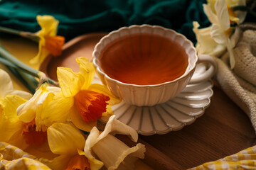 Close-up of a tea cup surrounded by yellow daffodils, wooden tray, green and cream fabrics, creating a warm and inviting springtime still life with natural light