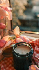 Close-up of a cozy windowsill scene with magnolia flowers in a glass jar, a black candle, and a cup of tea on a white tray with checkered fabric in warm evening light