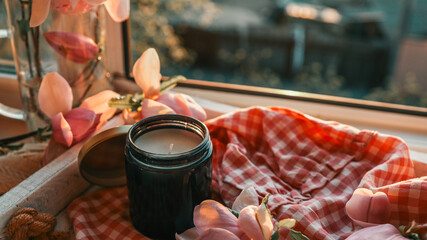 Close-up of a cozy windowsill scene with magnolia flowers in a glass jar, a black candle, and a cup of tea on a white tray with checkered fabric in warm evening light