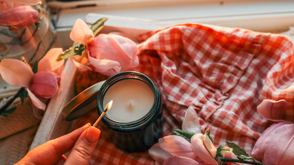Close-up of a hand lighting a black jar candle with a match, surrounded by soft pink flowers and a...